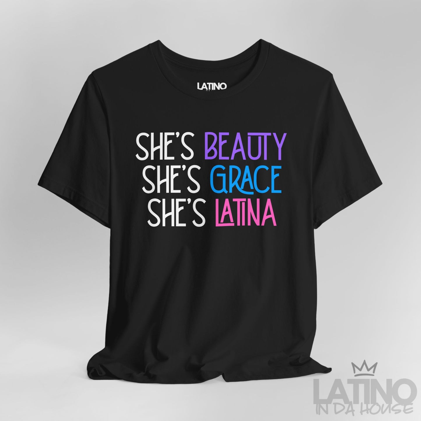 Close-up view of a black T-shirt with the phrase “She’s Beauty, She’s Grace, She’s Latina” in pink, blue, and purple font. Latino In Da House