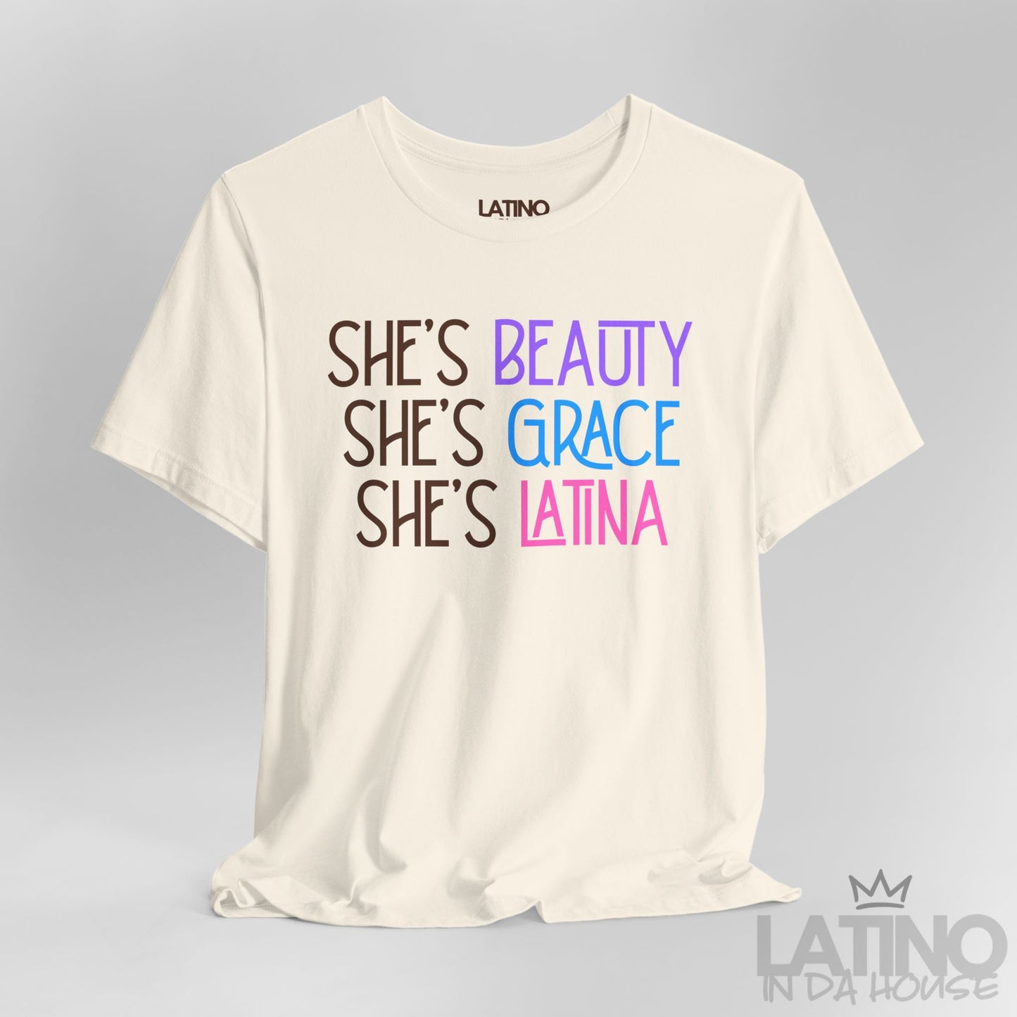 Close-up view of a natural color T-shirt with the phrase “She’s Beauty, She’s Grace, She’s Latina” in pink, blue, and purple font. Latino In Da House